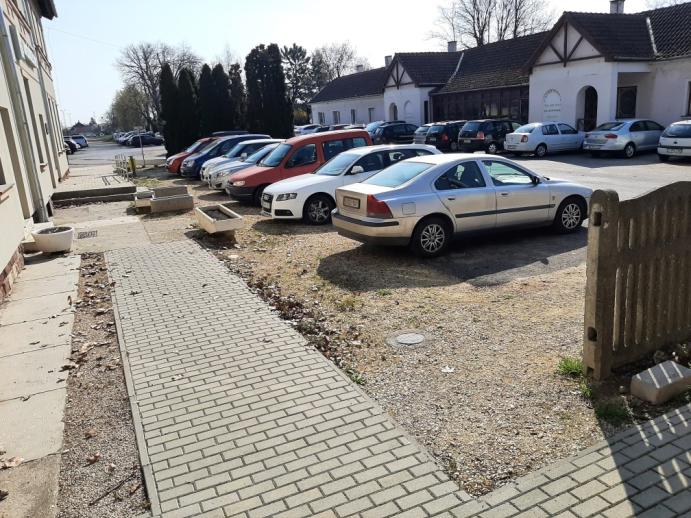 A Fertőszentmiklós vasútállomás előtti parkolóterület kerékpártárolóval, állomásépülettel és parkoló autókkal.