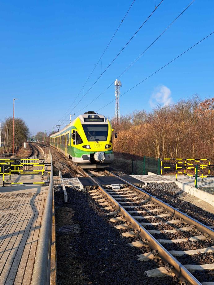 A képen egy GYSEV motorvonat közeledik egy gyalogos vasúti átjáróhoz. Az átjárót fekete-sárga korlátok és burkolt gyalogút jelöli a sínek között.