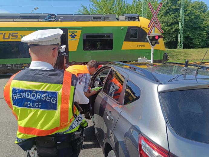 Rendőrök és vasúti szakemberek egy autó vezetőjével beszélnek egy vasúti átjáróban, miközben a háttérben egy GYSEV motorvonat halad el a lezárt átjáró mellett.