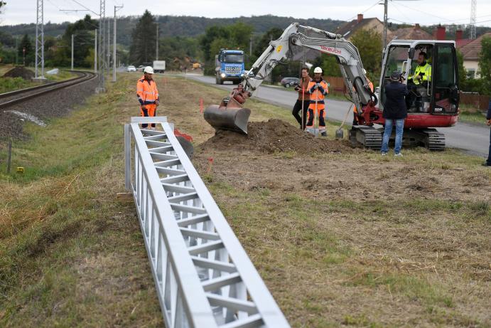 A vasúti pálya mellett munkások és egy munkagép dolgozik, az előtérben egy új felsővezetéki oszlop látható.
