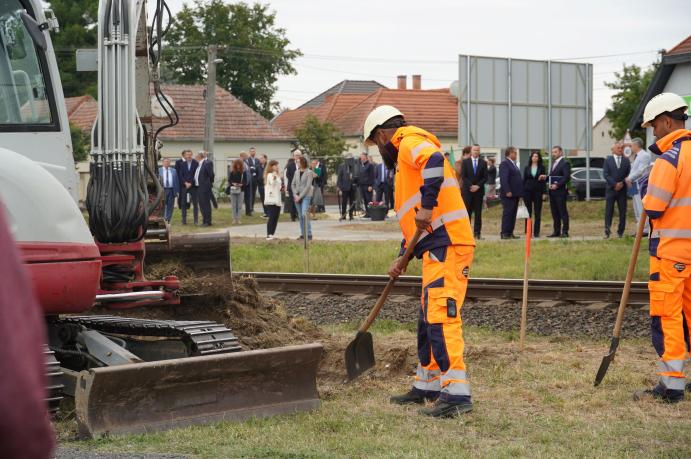 A vasúti pálya mellett munkások dolgoznak, a háttérben a nyitórendezvény résztvevői láthatók.